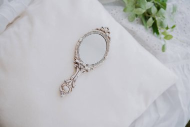 Yeşilliklerle çevrili yumuşak beyaz bir yastığın üzerinde antika bir el aynası yatıyor..