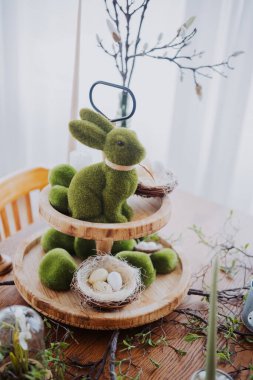 Bahar dekorasyonunda yuvalara yerleştirilmiş yeşil tavşanlı katmanlı bir tepsi ve yumurtalar yer alıyor..