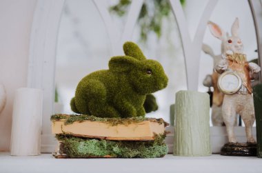 A green rabbit sits on a book with moss while candles and figurines decorate the space. clipart