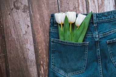 A pair of denim jeans with white tulips peeking from the pocket on a wooden surface. clipart