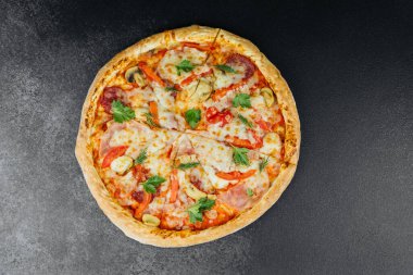 Peynirli, biberli, zeytinli ve jambonlu yuvarlak bir pizza koyu bir yüzeyde oturur..