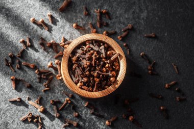 Karanlık bir zemin üzerine saçılmış baharatlarla karanfil dolu ahşap bir kase..