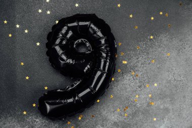 Dokuz numaraya benzeyen büyük siyah bir balon altın yıldızlı gri bir yüzeyde yatar..