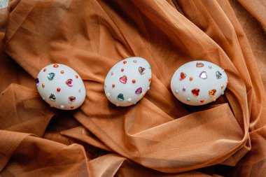 Üç eşsiz dekore edilmiş yumurta yumuşak bir kumaş yüzeyinde duran renkli mücevherler..