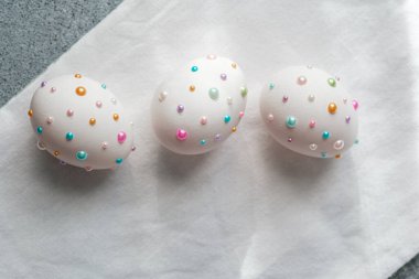 Gri bir yüzeyde renkli incilerle süslenmiş üç beyaz yumurta neşeli bir görünüm yaratır..
