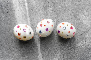 Renkli kalpler ve noktalarla süslenmiş üç beyaz yumurta gri bir arkaplanda oturur..