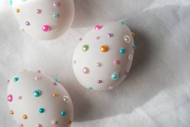 Yumuşak bir yüzeyde duran renkli boncuklarla süslenmiş üç beyaz yumurta..