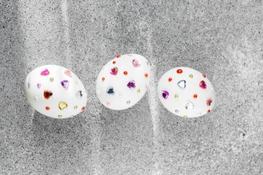 Canlı mücevherlerle süslenmiş üç beyaz yumurta gri bir yüzey üzerinde sergilenir..