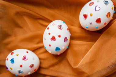 Turuncu kumaş üzerinde canlı kalp şeklinde mücevherlerle süslenmiş üç beyaz yumurta..