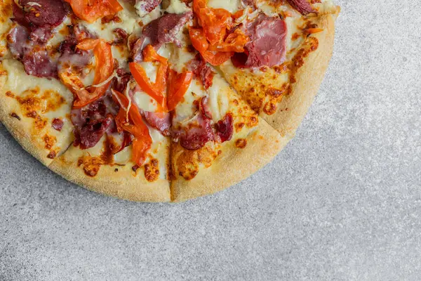 Taze pişmiş et ve sebzelerle kaplı bir pizza gri bir yüzeyde duruyor..