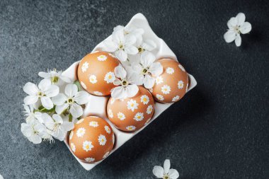 Dekoratif yumurtalar, karanlık bir yüzey üzerinde narin beyaz çiçeklerle çevrili bir kapta otururlar..