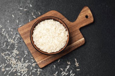 Beyaz pirinçle dolu ahşap bir kase düz bir kesme tahtasının üzerinde duruyor, dağınık taneciklerle..