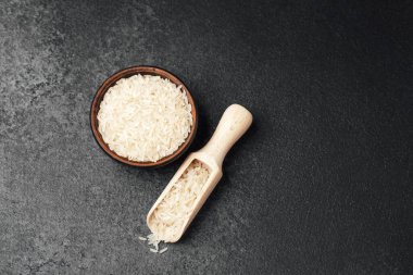 Koyu bir yüzeyde ahşap bir kepçenin yanında pişmemiş beyaz pirinçle dolu bir kase..