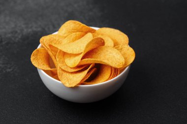 Beyaz bir kase dolusu altın çıtır patates cipsi karanlık bir masada duruyor..