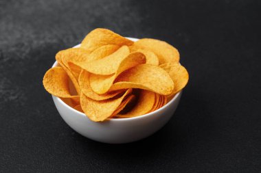 Beyaz bir kase dolusu altın çıtır patates cipsi karanlık bir masada duruyor..