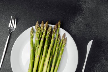 Yeşil kuşkonmaz sapları beyaz bir tabağa yerleştirilmiş, yanında da çatal bıçak takımı var..