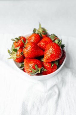 Parlak kırmızı çileklerle dolu bir kase yumuşak beyaz bir kumaşın üzerinde duruyor..