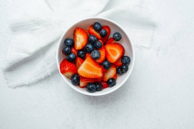 Dilimlenmiş çilek dolu bir kase ve tüm yaban mersini açık bir yüzeyde bulunur..