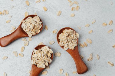 Üç ahşap kaşık yulafları gri bir zemin üzerinde tutar, tahıllar etrafa saçılmıştır..