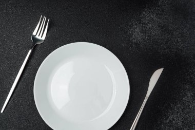 Koyu dokuda beyaz bir tabak ve gümüş çatal bıçak içeren minimalist bir düzen..