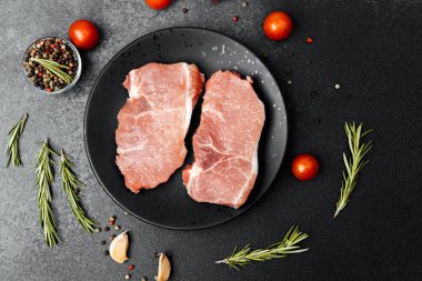 Çiğ domuz pirzolası siyah bir tezgahta otlar, baharatlar ve domateslerle sergilenir..