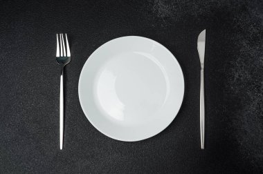 Siyah bir yüzeye, yanına çatal ve bıçak yerleştirilmiş beyaz bir plaka yerleştirilir..
