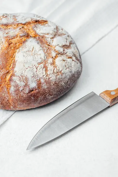 Yuvarlak bir somun ekmek, sade bir mutfak yüzeyinde bıçağın yanında oturur..