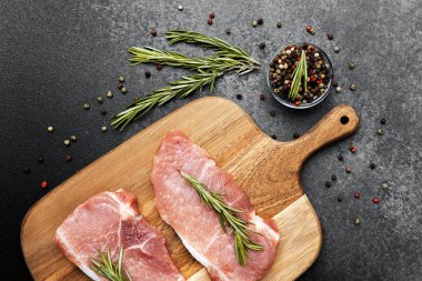 İki çiğ et parçası bir kesme tahtasının üzerinde otlar ve baharatların yanında duruyor..