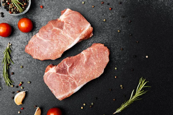 İki çiğ domuz pirzolası, baharatlar, sarımsak ve vişneli domateslerle çevrili..