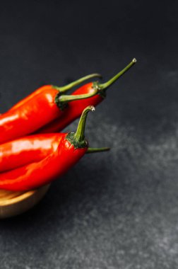 Üç taze kırmızı biber, koyu dokulu bir yüzey üzerinde küçük bir ahşap kasede dinleniyor..