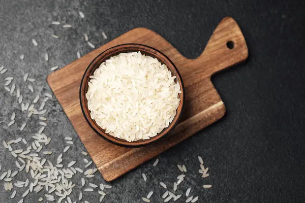 Beyaz pirinçle dolu ahşap bir kase düz bir kesme tahtasının üzerinde duruyor, dağınık taneciklerle..