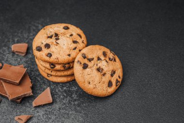 Bir yığın çikolata parçalı kurabiye koyu bir yüzeyde kırık çikolata parçalarının yanında duruyor..