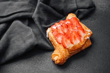 Dilimlenmiş taze çilek ve kremayla kaplı yumuşak bir hamur işi siyah bir yüzeye oturur..