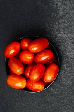 Parlak vişneli domatesler, dokulu bir yüzeyde siyah bir kasede nemli bir şekilde parlarlar..