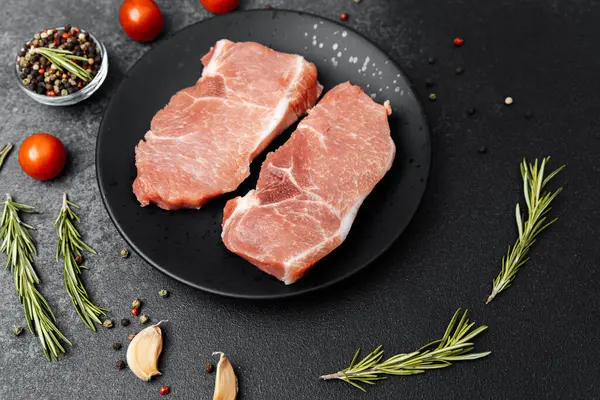 İki çiğ domuz pirzolası siyah bir tabağa konmuş. Etrafı ot ve baharatla çevrili..