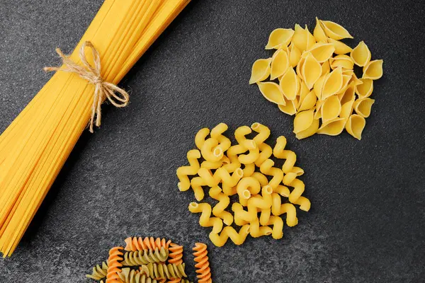 Yakınlarda bir demet spagetti ile koyu bir yüzeyde çeşitli makarna tipleri sergileniyor..