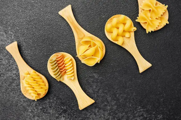 Tahta bir yüzey üzerinde ahşap kaşıklarla dizilmiş çeşitli pişmemiş makarna şekilleri.