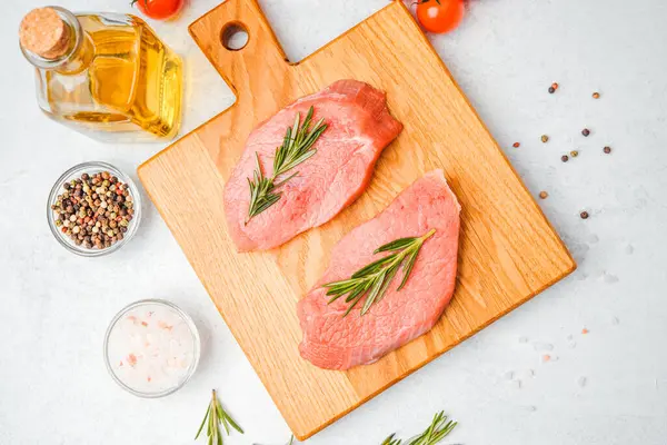 Domates, yağ, tuz ve biberiye ile çevrili ahşap kesim tahtasıyla çiğ dana bifteği.