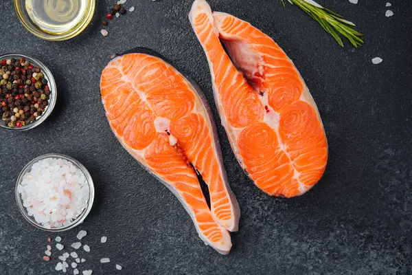 İki canlı somon filetosu karanlık bir yüzeyde otlar ve baharatlarla sıralanmış..