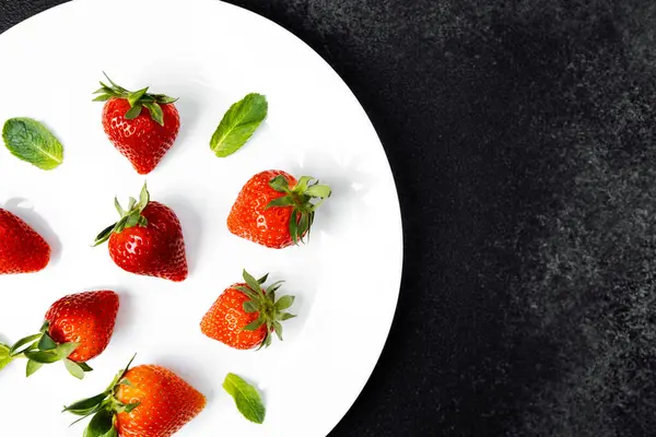 Garnitür olarak yeşil nane yapraklı beyaz bir tabakta sergilenen sulu çilekler..