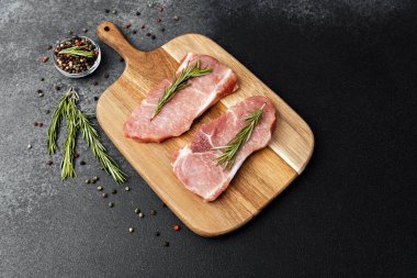 İki domuz pirzolası ahşap bir tahtaya konur ve biberiye ve biber etrafa saçılır..