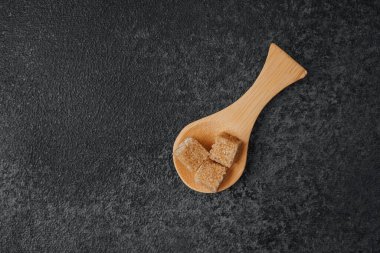 Esmer şeker küpleri, ahşap bir kaşığın üzerinde, koyu renkli bir yüzey üzerinde dinleniyor..