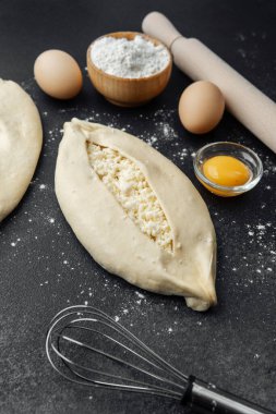 İki çiğ khachapuri ekmeği, içinde fırın malzemeleri olan siyah un kaplı bir masada.