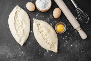 İki çiğ khachapuri ekmeği, içinde fırın malzemeleri olan siyah un kaplı bir masada.