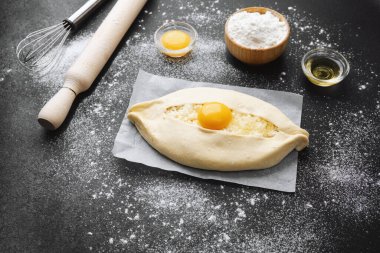 Çiğ khachapuri, yumurta sarısı ve parşömen üzerinde peynir ve siyah arka planda un.