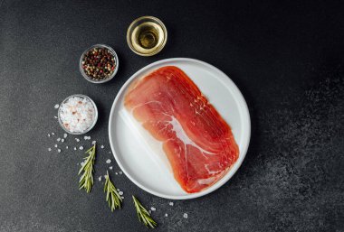 Beyaz tabakta dilimlenmiş jambon ve siyah arka planda baharatlar.