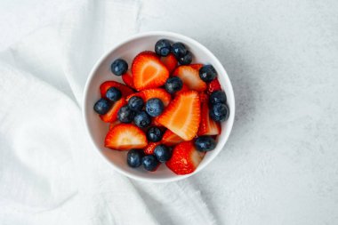 Dilimlenmiş çilek dolu bir kase ve tüm yaban mersini açık bir yüzeyde bulunur..