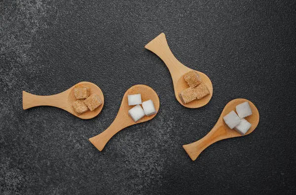 Koyu bir yüzeyde küçük tahta kaşıklarda sıralanmış farklı şeker türleri.