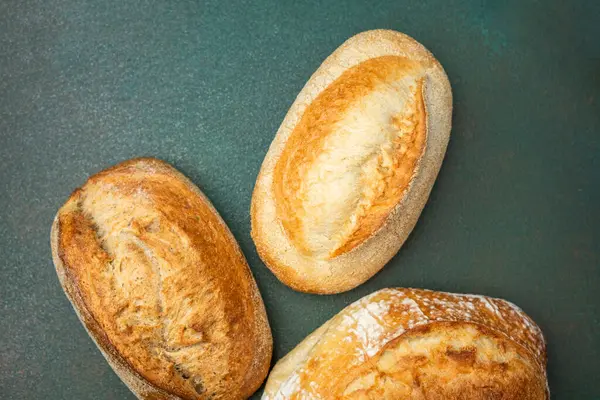 Koyu yeşil bir yüzey üzerinde üç somun ekmek sergileniyor, dokularını renklendiriyor..