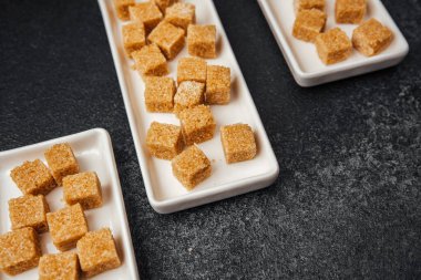 Kahverengi şeker küpleri beyaz tabakalarda sergilenir ve koyu bir yüzey üzerinde dinlenir. Kontrastlık oluşturur.